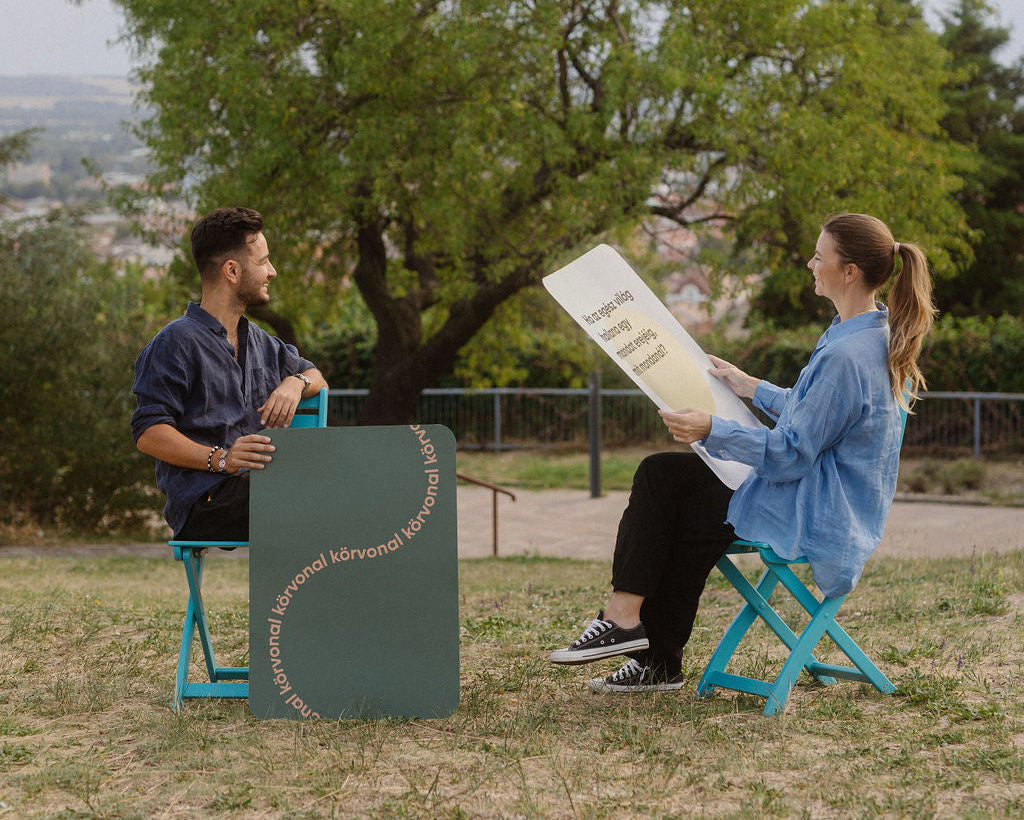 Két ember beszélget egy park fái alatt, egy nagy körvonal kártyával